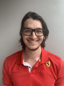 Connor Kapahi standing in front of a white wall in a red polo shirt and black glasses